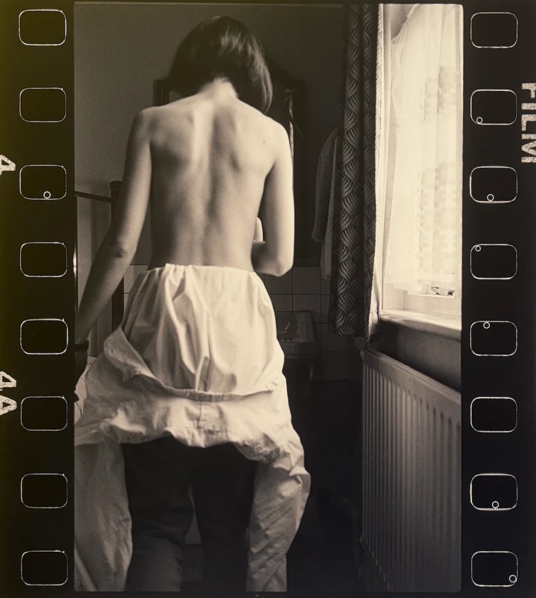 Jyll Bradley, Self-Portrait, bareback 1987–2023: black-and-white photograph framed by film-strip edges, a woman seen from behind in a bedroom beside a window with lace curtains